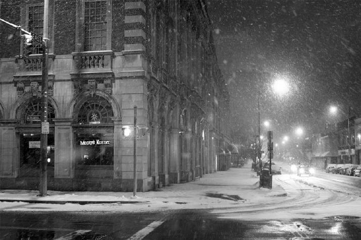 Snow falls gently during the night on a deserted New York Street lit by glowing street lamps. 