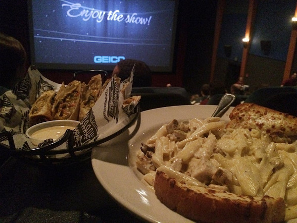 food forgeground with theater screen in the background "Enjoy the show!"; food on the right is a pasta dish with lots of cheese and cheesy bread; food on the left might be tacos