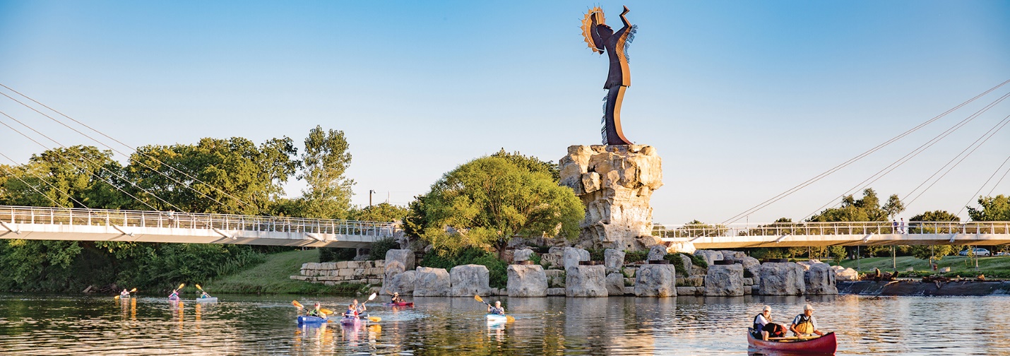 wide shot of the sculpture at center with the river in the foreground; people in canoes on the river