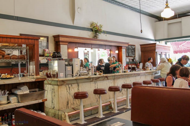 old fashioned looking soda shop; marble counter, stools along it with a mirror behind it; booths in the foreground