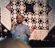 Armin Shimerman sitting in front of a quilt which is hanging on the wall. The picture isn't large enough to discern many details, but it appears to be mostly white, black, and grey.