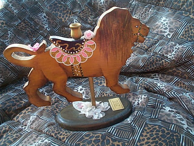 carousel lion; it's  a candle holder; the lion is on a dowel mounted on an oval wooden base; the lion is painted wood, wearing a bridle around its mouth; saddle is black with gold trip and pink ruffles; pink roses where the saddle knob would be and on the lion's tail, which doubles back to the lion's backside
